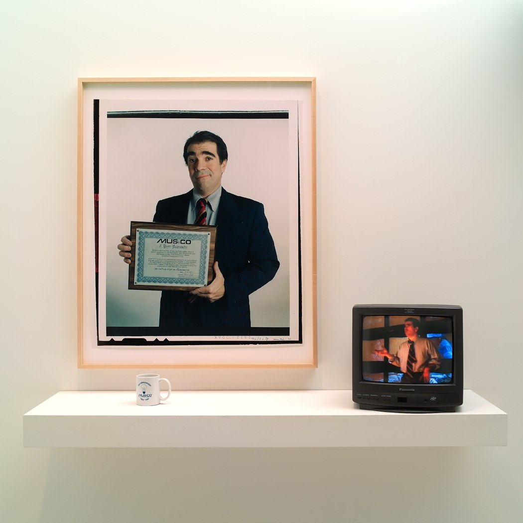 video still shows man gesturing in front of wall of video monitors